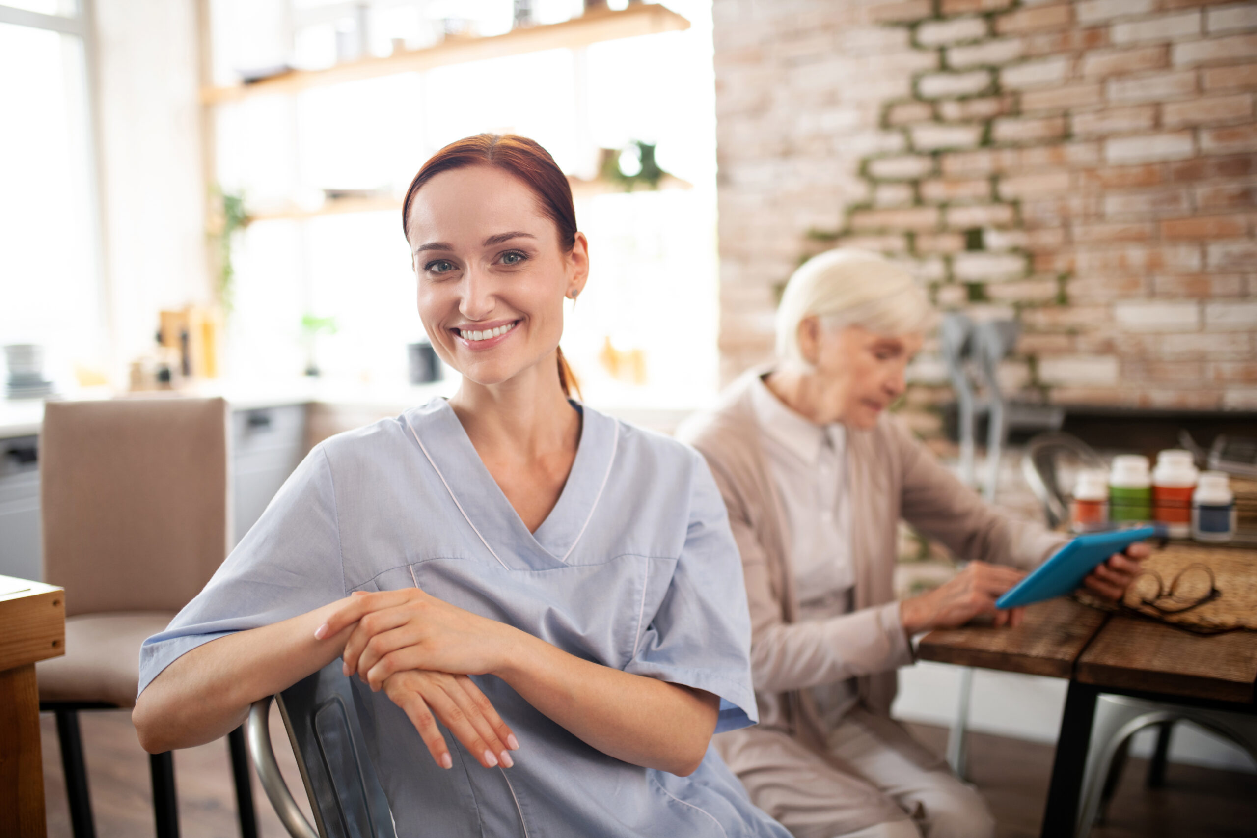 Smiling caregiver in a blue uniform assisting an elderly woman at home. Home care jobs in Philadelphia offer competitive pay and benefits.