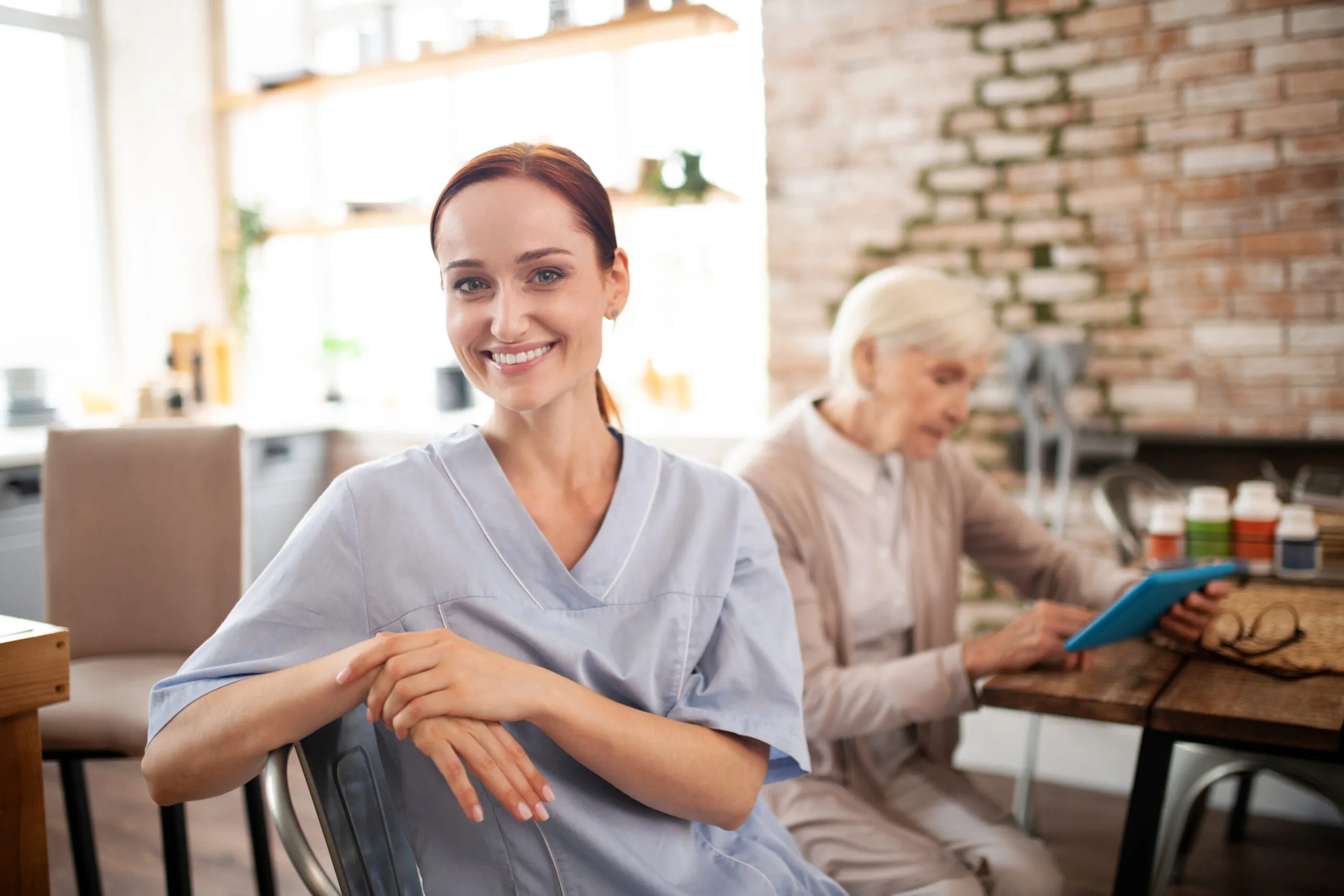 Smiling caregiver in a blue uniform assisting an elderly woman at home. Home care jobs in Philadelphia offer competitive pay and benefits.